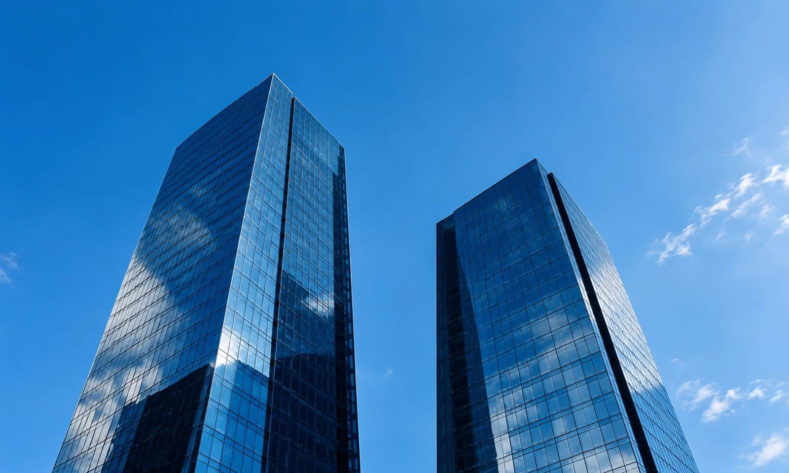 Two modern glass office towers side by side against a clear blue sky, one taller than the other.