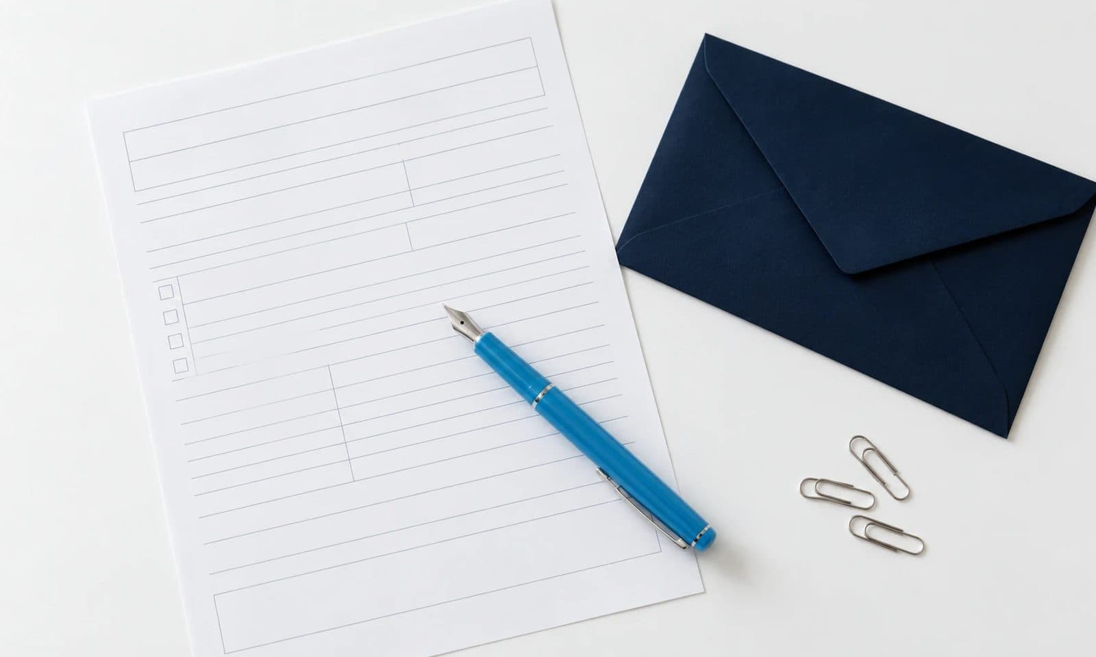 A blank tax form with a blue fountain pen beside a navy envelope and paper clips on a white desk.